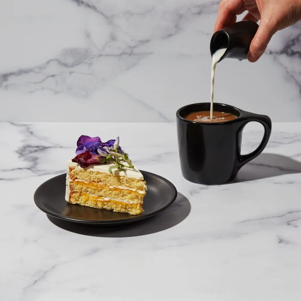 Coffee being poured into a black notNeutral Lino mug beside a slice of cake